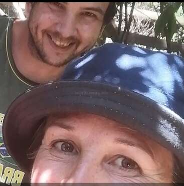 Two people smiling outdoors, one wearing a blue bucket hat in the foreground, with sunlight casting shadows on their faces, creating a warm and friendly atmosphere.