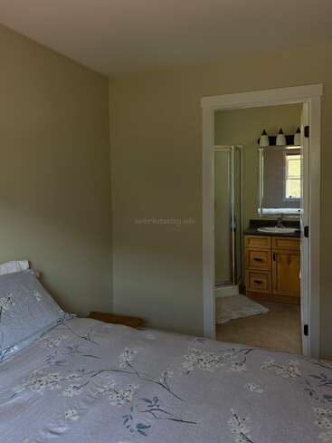 Un acogedor dormitorio con una colcha de motivos florales, paredes claras y un cuarto de baño con armarios de madera, una pequeña ventana y una moderna iluminación sobre el espejo.