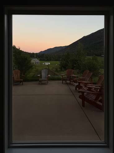 Un gran ventanal enmarca un patio con sillas de madera vacías, con vistas a un apacible paisaje rural, una casa lejana, campos verdes y montañas bajo un cielo en tonos pastel al atardecer.