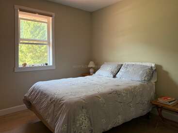 Un dormitorio pequeño y acogedor con paredes de color claro, una cama cubierta con ropa de cama de flores, una mesilla de noche de madera, libros, una lámpara y una ventana iluminada por el sol.