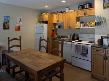 Esta acogedora cocina cuenta con armarios de madera, una mesa de comedor de madera con cuatro sillas, electrodomésticos blancos, cuchillos montados en la pared, colorido arte mural y utensilios de cocina de uso cotidiano.