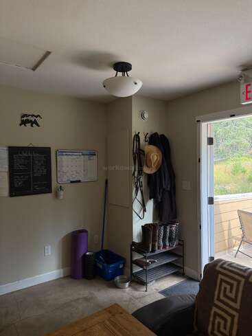 Una pequeña entrada con un calendario de pared, una pizarra, abrigos, sombreros, botas, una esterilla de yoga, utensilios de limpieza y una puerta abierta que da a una zona de césped.