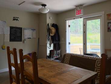 Una acogedora habitación con una mesa de madera, sillas, un sofá, abrigos y sombreros colgados, botas, una pizarra, un calendario y una puerta de cristal que da al exterior.
