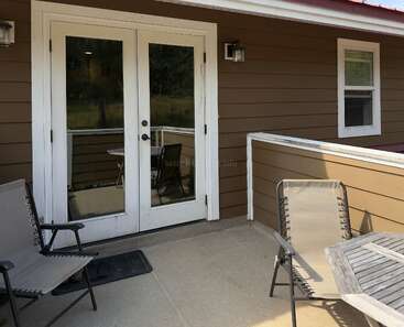 Esta imagen muestra un patio con dos tumbonas, una mesa de madera y puertas francesas que dan al interior. La pared exterior es marrón con ribetes blancos.