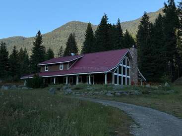 Una casa rústica con tejado rojo se asienta entre altos pinos y paisajes de montaña. Se ven grandes ventanales, una chimenea de piedra y un sinuoso camino de grava.
