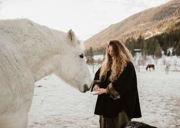 Una mujer con el pelo largo está de pie en un campo nevado, sonriendo mientras da de comer a mano a un gran caballo blanco. Al fondo se ven montañas boscosas.