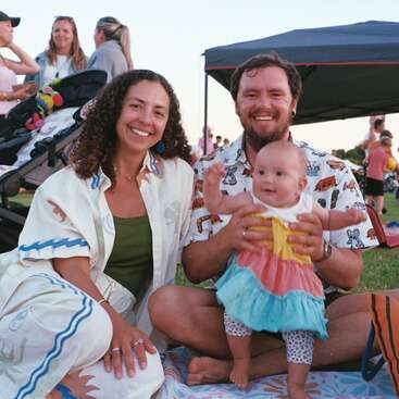 Une joyeuse famille de trois personnes est assise sur une couverture de pique-nique en plein air. Les parents sourient, le visage rayonnant de bonheur, tandis qu'ils tiennent leur bébé dans leurs bras. À l'arrière-plan, d'autres personnes discutent entre elles.