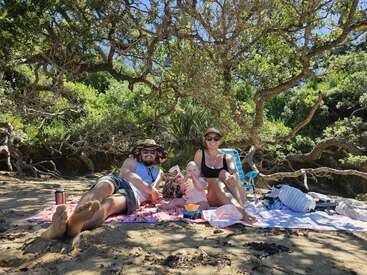 Eine Familie entspannt gemeinsam bei einem Picknick am Sandstrand im Schatten großer Bäume und genießt die Sonne, kleine Snacks und die schöne Zeit draußen in der Natur.