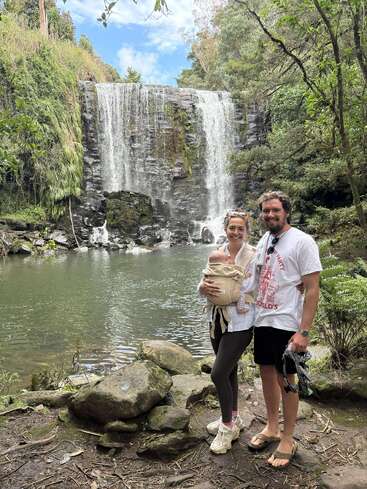 Ein lächelndes Paar posiert vor einem wunderschönen Wasserfall, umgeben von üppigem Grün. Die Frau trägt ein Baby in einem Tragetuch. Naturszene, im Freien.