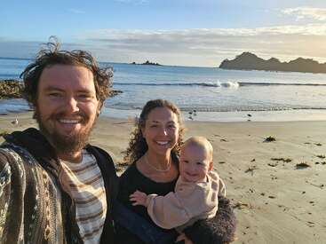Eine lächelnde dreiköpfige Familie genießt einen sonnigen Tag am Strand; im Hintergrund sind unter blauem Himmel Wellen, Möwen und felsige Inseln zu sehen.