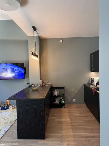 A modern kitchen with black cabinets, wood floor, and a dark island. Pendant lights hang above. A TV displays Earth, and children’s toys are visible.