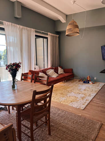 A cozy living room features a red sofa, light curtains, wooden dining table, playful children's toys, soft rugs, a woven pendant lamp, and fresh flowers.