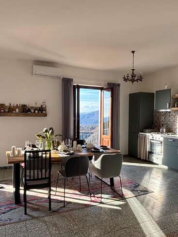 A sunlit dining room with a set table, modern chairs, open window revealing mountains, cozy kitchen, tiled floor, decorative chandelier, and a shelf of bottles.