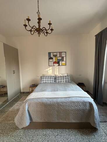 A cozy bedroom with a neatly made bed, checkered pillows, modern abstract artwork, mirrored wardrobe, vintage chandelier, side tables, and a window with gray curtains.