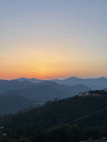 The image depicts a serene sunset over a mountain range, with a small house on the right side, surrounded by lush trees and power lines, set against a vibrant orange and yellow sky.