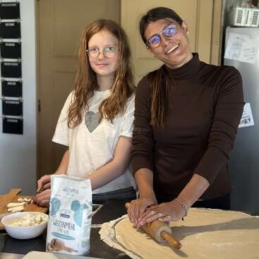 A woman and a girl are happily preparing food together in a kitchen, rolling dough and chopping ingredients, creating a warm, family cooking atmosphere.