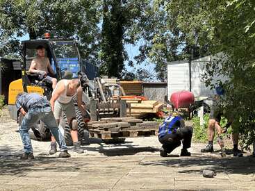Cinq hommes travaillent à l'extérieur avec des pavés, à l'aide d'une petite chargeuse et d'outils, par temps ensoleillé, entourés de verdure, de hangars et de matériaux de construction, dans un esprit de travail d'équipe évident.