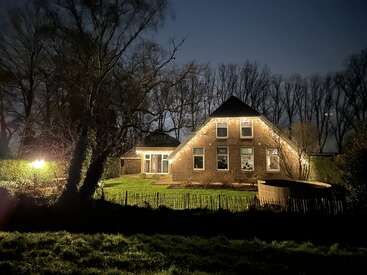 Une maison en briques confortable est chaleureusement éclairée la nuit par des guirlandes lumineuses, entourée d'arbres et de verdure, créant une atmosphère paisible et accueillante à la campagne.