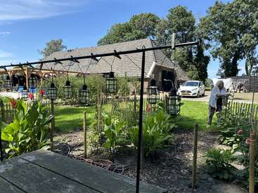 Une scène paisible dans un jardin avec des lanternes suspendues, des plantes vertes luxuriantes, une maison rustique en bois, une voiture blanche garée et une femme âgée qui s'occupe des plantes.