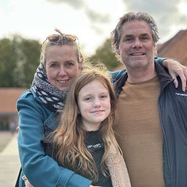 Une famille de trois personnes sourit en posant à l'extérieur. La femme porte un foulard et des lunettes de soleil, l'homme une veste, et la jeune fille se tient devant, l'air joyeux.