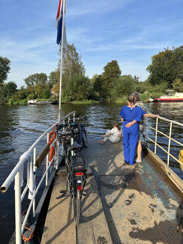 Deux vélos sont garés sur un ferry traversant une rivière. Trois personnes profitent de la vue panoramique, avec une végétation luxuriante, des bateaux et un ciel bleu en arrière-plan.