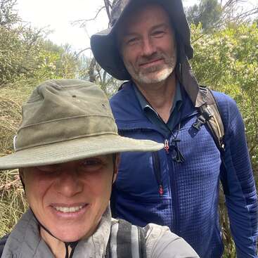 Zwei Menschen lächeln im Freien, tragen breitkrempige Hüte und Wanderausrüstung. Umgeben von grünem Laub und Bäumen scheinen sie auf einem Natur- oder Wanderabenteuer zu sein.