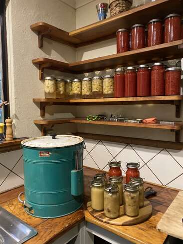 In einer gemütlichen Küchenecke stehen Regale voller hausgemachter Konfitüren in Gläsern, ein großer Vintage-Einmachkessel und frisch verschlossene Gläser mit Soße und Obst.