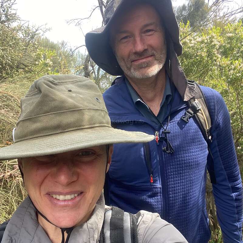 Zwei Menschen lächeln im Freien, tragen breitkrempige Hüte und Wanderausrüstung. Umgeben von grünem Laub und Bäumen scheinen sie auf einem Natur- oder Wanderabenteuer zu sein.