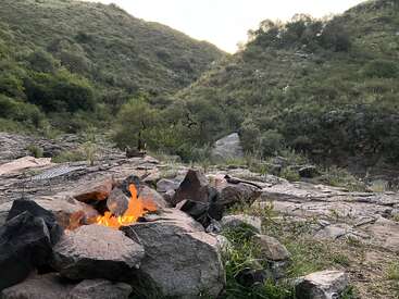 A small campfire surrounded by rocks burns brightly on rocky ground. Green hills and trees fill the background, creating a peaceful outdoor wilderness setting.