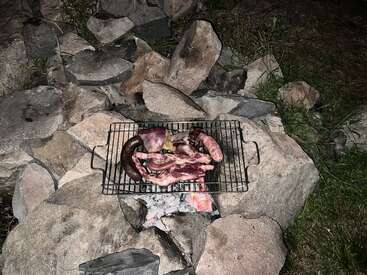 In the image, various meats are grilling on a metal rack over glowing embers, surrounded by large stones outdoors at night, creating a rustic barbecue setting.