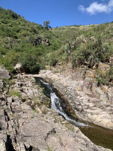 A small waterfall flows over rocky terrain surrounded by lush green vegetation. Hills rise in the background under a clear blue sky with a few clouds.
