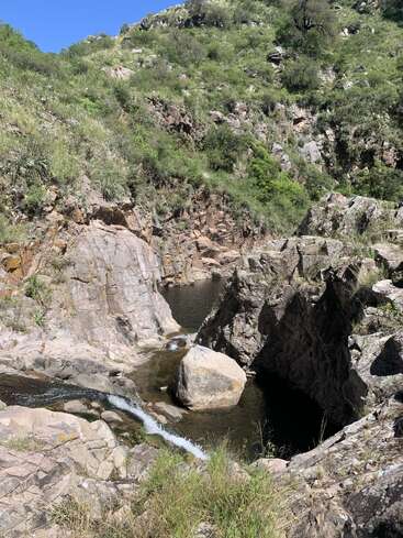 A rocky canyon with a small stream of water flows between boulders, surrounded by lush green vegetation and steep hills under a clear blue sky.