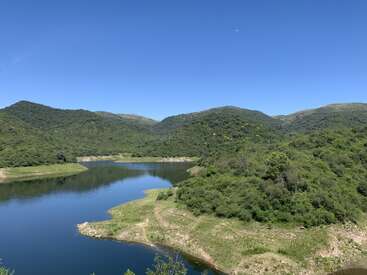 A serene scene of green hills surrounds a calm, winding river under a clear blue sky. Lush vegetation covers the landscape, evoking peaceful natural beauty.