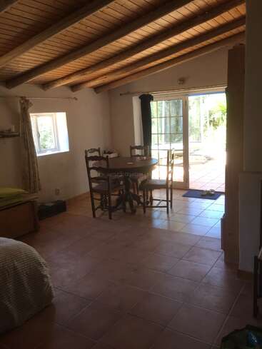 Este quarto aconchegante tem teto com vigas de madeira, piso de azulejo, mesa redonda com cadeiras, janelas pequenas, cama e portas de vidro que dão para um pátio ensolarado.