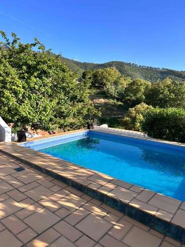 Uma piscina azul cintilante fica em um pátio de azulejos, cercada por árvores verdejantes, com vista para colinas onduladas sob um céu azul claro e brilhante. Tranquilo.