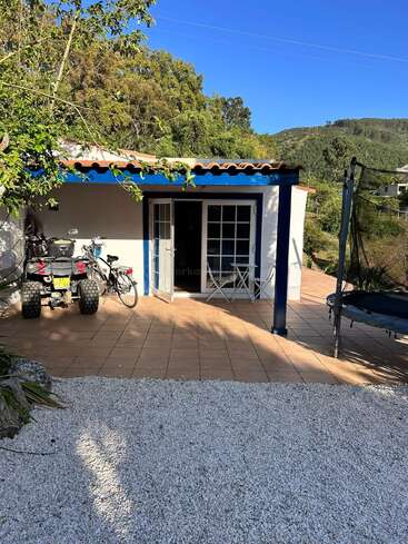 Uma casinha branca com detalhes azuis tem um pátio de azulejos, mesa de jardim, cadeiras, bicicleta, quadriciclo, cama elástica, quintal de cascalho, cercada por árvores e colinas.