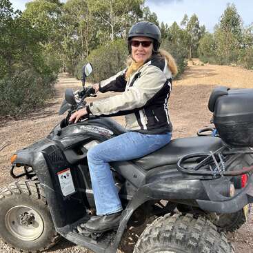 Une femme portant un casque, des lunettes de soleil, une veste et un jean est assise sur un quad noir, sur un chemin de terre entouré d'arbres et de verdure.