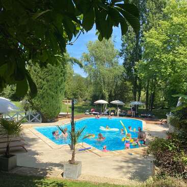 Kinder und Erwachsene genießen einen sonnigen Tag beim Schwimmen in einem leuchtend blauen Pool, umgeben von üppig grünen Bäumen, Rasen, Liegestühlen und weißen Sonnenschirmen. Entspannende Atmosphäre.