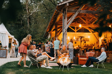 Eine Gruppe von Menschen genießt ein gemütliches Beisammensein im Freien. Einige sitzen um eine Feuerstelle herum, andere mischen sich unter einen warm beleuchteten, rustikalen Holzunterstand und schaffen eine lebendige Atmosphäre.