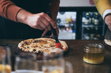 Eine Person bestreicht eine frisch gebackene Pizzakruste mit einem roten Backpinsel mit Olivenöl, während eine andere Person in der Nähe das Essen zubereitet. Gläser und ein Einmachglas sind zu sehen.