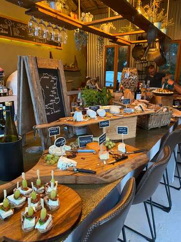 Eine rustikale französische Käsebar mit verschiedenen Käsesorten, Trauben, Brot, Wein, beschrifteten Platten, gemütlicher Beleuchtung, hängenden Gläsern und Vintage-Dekor schafft eine warme, einladende Atmosphäre.