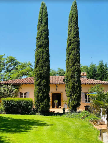 Ein charmantes Haus mit Ziegeldach und hellen Wänden steht hinter zwei hohen Zypressen, umgeben von üppigem Grün und einem gepflegten Rasen.
