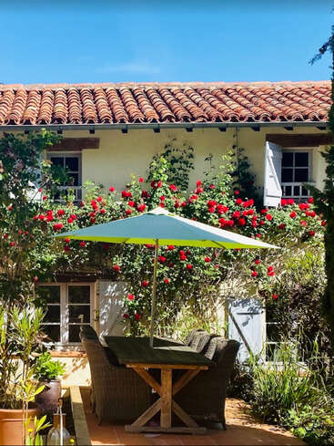 Eine gemütliche Terrassenszene mit einem Holztisch, Korbstühlen und einem Sonnenschirm, umgeben von leuchtend roten Rosen, vor einem charmanten Häuschen mit Terrakotta-Dachziegeln.