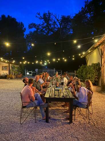 Eine Gruppe von Menschen genießt in der Abenddämmerung ein Abendessen im Freien unter Lichterketten, versammelt um einen langen Holztisch, umgeben von Bäumen und rustikalen Gebäuden.