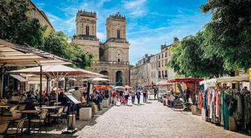Eine lebhafte Marktszene unter freiem Himmel mit Menschen, die spazieren gehen, unter Sonnenschirmen essen und an bunten Ständen stöbern. Historische Steingebäude und Bäume umgeben den lebhaften, sonnenbeschienenen Platz.