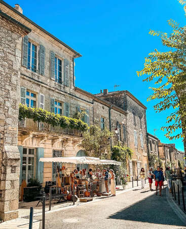 Eine charmante Straßenszene in einem malerischen europäischen Dorf mit steinernen Gebäuden, offenen Marktständen, Menschen, die einkaufen, und einem klaren blauen Himmel unter warmem Sonnenschein. Angenehme Atmosphäre.