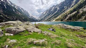 Un magnifique paysage de montagne composé de reliefs rocheux, d'une pelouse verdoyante, d'un lac paisible et de sommets enneigés. Deux personnes se détendent sur l'herbe, profitant de la nature.