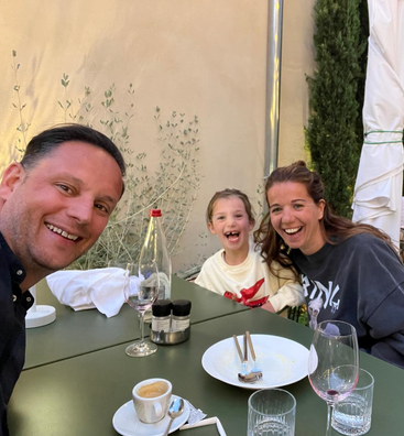 Une famille heureuse de trois personnes est assise à une table verte en plein air, partageant des rires après avoir terminé un repas, entourée d'assiettes, de verres et de cafés vides.