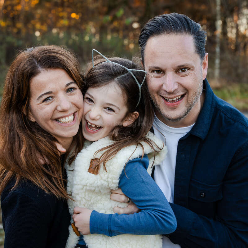 Eine glückliche dreiköpfige Familie posiert lächelnd und lachend im Freien. Das junge Mädchen trägt ein Stirnband mit Katzenohren und wird von ihren lächelnden Eltern in der Herbstlandschaft umarmt.