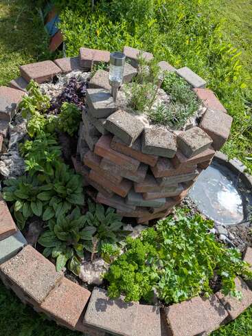 This image shows a spiral-shaped herb garden made with bricks, various herbs growing inside, solar garden light on top, surrounded by lush green grass and plants.